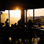Travelers sitting in an airport terminal at sunset, silhouetted against the backdrop of an airplane.